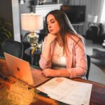 Georgia sitting at a desk in front of her laptop and paperwork looking at the screen with her hand on her chin