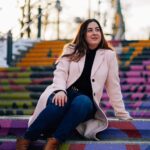 Georgia is sitting on an outdoor painted staircase in a pink coat